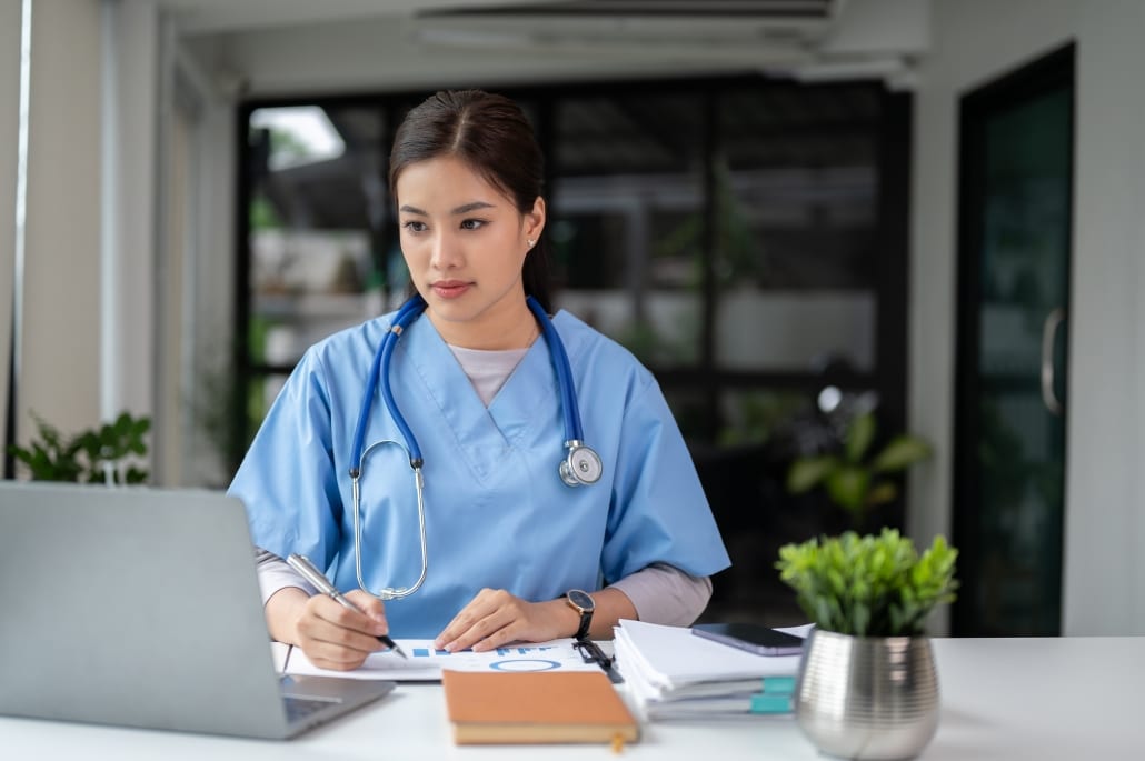Asian nurse in scrubs taking notes and using a laptop